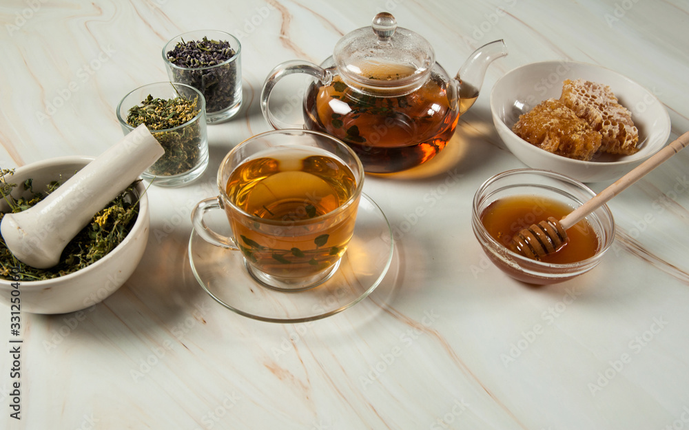 Healthy and delicious herbal tea with honey. A teapot, tea in a glass cup, sweet golden honey and a set of dried herbs in a porcelain mortar with a pestle close-up on a pastel green marble background. Healthy and delicious herbal tea with honey. A teapot, tea in a glass cup, sweet golden honey and a set of dried herbs in a porcelain mortar with a pestle close-up on a pastel green marble background.