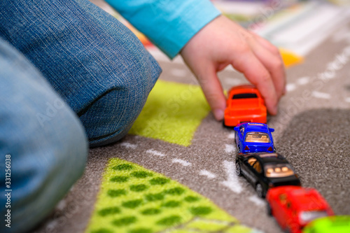 Close-up of five year old boy playing and lining up toy cars on a playing mat with roads. The cars have vivid colors and the boy is dressed in blue jeans and a blue shirt. He is holding a red pickup t