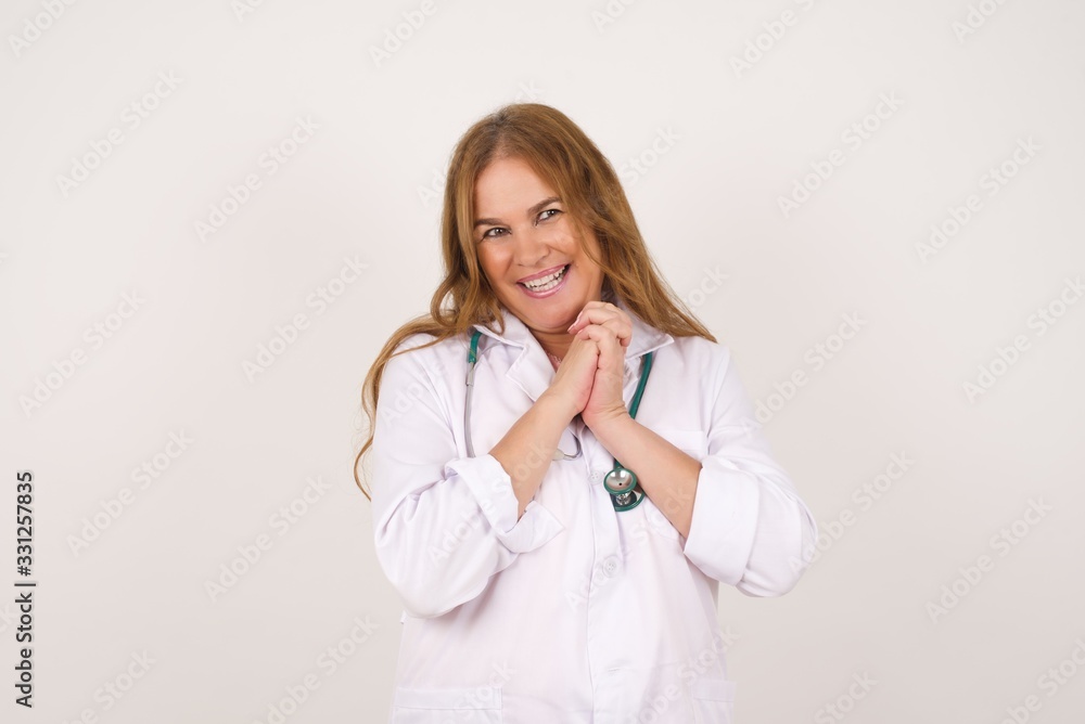 Dreamy young doctor woman with pleasant expression, closes eyes, keeps hands crossed near face, thinks about something pleasant, poses against gray background. Dreamy young doctor woman with pleasant expression, closes eyes, keeps hands crossed near face, thinks about something pleasant, poses against gray background.