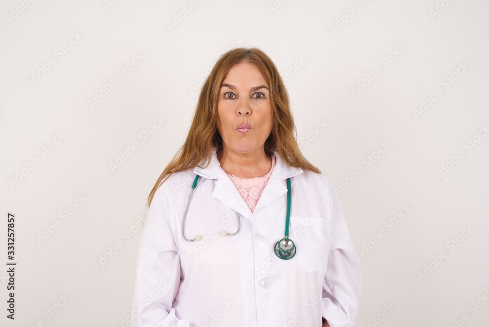 Attractive doctor female rounds lips, wears medical uniform, looks directly into camera, expresses her satisfaction, isolated over gray background. Attractive doctor female rounds lips, wears medical uniform, looks directly into camera, expresses her satisfaction, isolated over gray background.