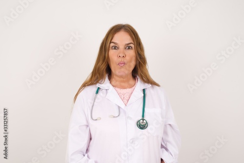 Attractive doctor female rounds lips  wears medical uniform  looks directly into camera  expresses her satisfaction  isolated over gray background.