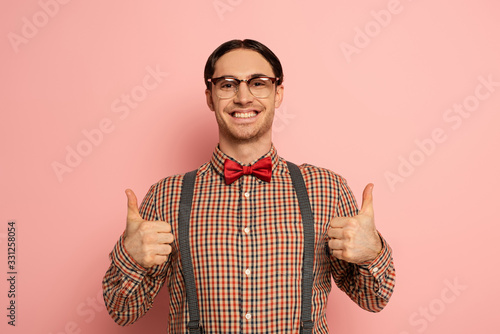 cheerful male nerd in eyeglasses showing thumbs up on pink