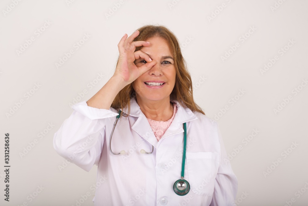 Beautiful young doctor female with happy face smiling doing ok sign with hand on eye looking through finger. Wearing medical uniform standing against gray wall. Beautiful young doctor female with happy face smiling doing ok sign with hand on eye looking through finger. Wearing medical uniform standing against gray wall.