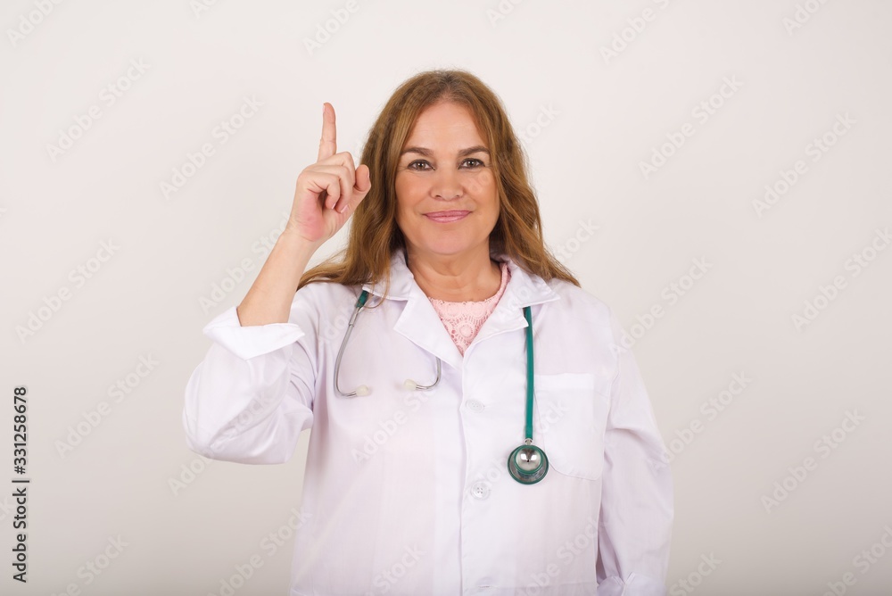 Young caucasian doctor woman wearing medical uniform, standing against gray wall showing and pointing up with fingers number one while smiling confident and happy.