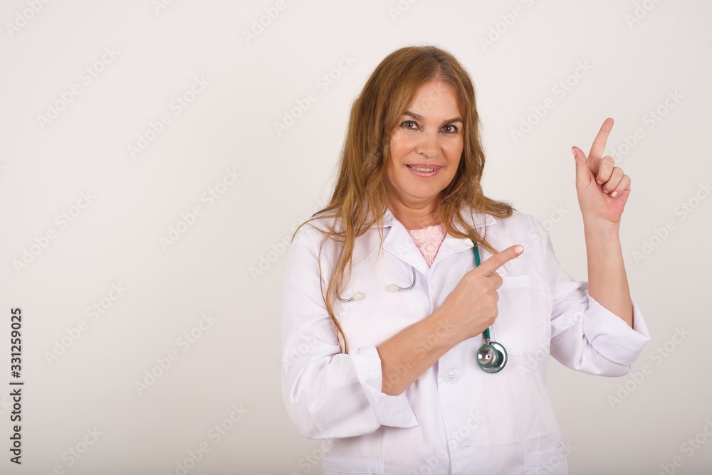 Pretty young doctor woman with positive expression, wearing medical uniform, indicates with fore finger at blank copy space for your promotional text or advertisement. Pretty young doctor woman with positive expression, wearing medical uniform, indicates with fore finger at blank copy space for your promotional text or advertisement.
