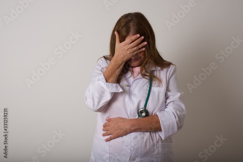 Indoor portrait of beautiful doctor woman, wearing medical uniform, making facepalm gesture while smiling, standing over gray background amazed with stupid situation. photo