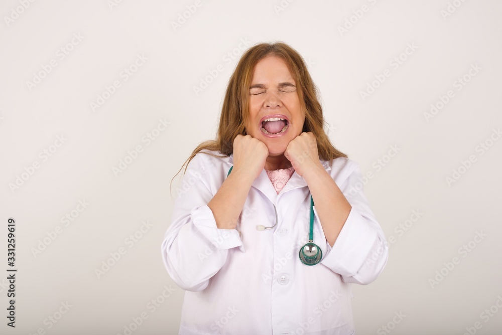 Portrait of young cute doctor woman being overwhelmed with emotions, expressing excitement and happiness with closed eyes and hands near face while smiling broadly over gray background. Portrait of young cute doctor woman being overwhelmed with emotions, expressing excitement and happiness with closed eyes and hands near face while smiling broadly over gray background.
