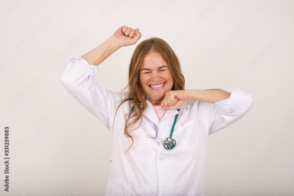 Attractive young caucasian doctor woman celebrating a victory punching the air with her fists and a beaming toothy smile over a gray studio background with copy space Attractive young caucasian doctor woman celebrating a victory punching the air with her fists and a beaming toothy smile over a gray studio background with copy space
