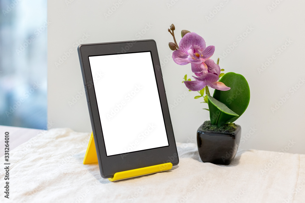 Modern gray electronic book or tablet with a blank mockup screen on a yellow stand next to an artificial pink orchid on the kitchen table with a linen tablecloth by the window Modern gray electronic book or tablet with a blank mockup screen on a yellow stand next to an artificial pink orchid on the kitchen table with a linen tablecloth by the window