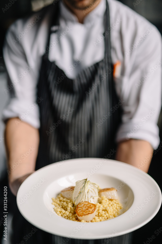 Cooker holding a plate with ptitim pasta, grilled scallops, cream sauce and dried onion slices Cooker holding a plate with ptitim pasta, grilled scallops, cream sauce and dried onion slices