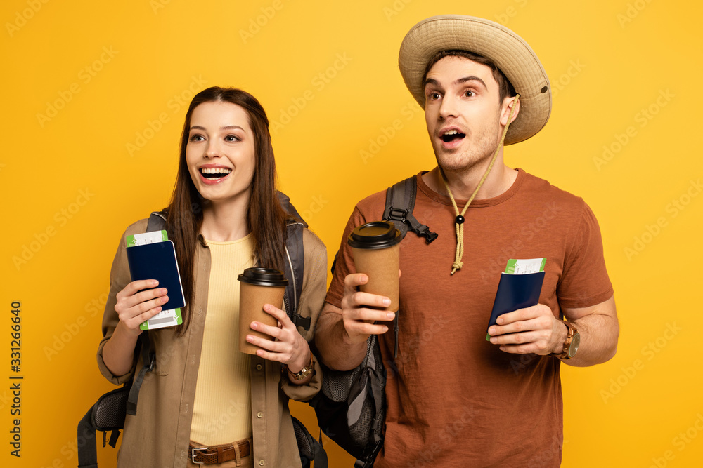 couple of excited travelers with backpacks holding passports, air tickets and coffee to go on yellow couple of excited travelers with backpacks holding passports, air tickets and coffee to go on yellow