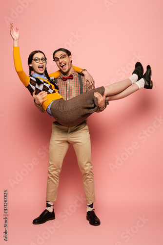excited nerd in eyeglasses holding girlfriend on hands on pink