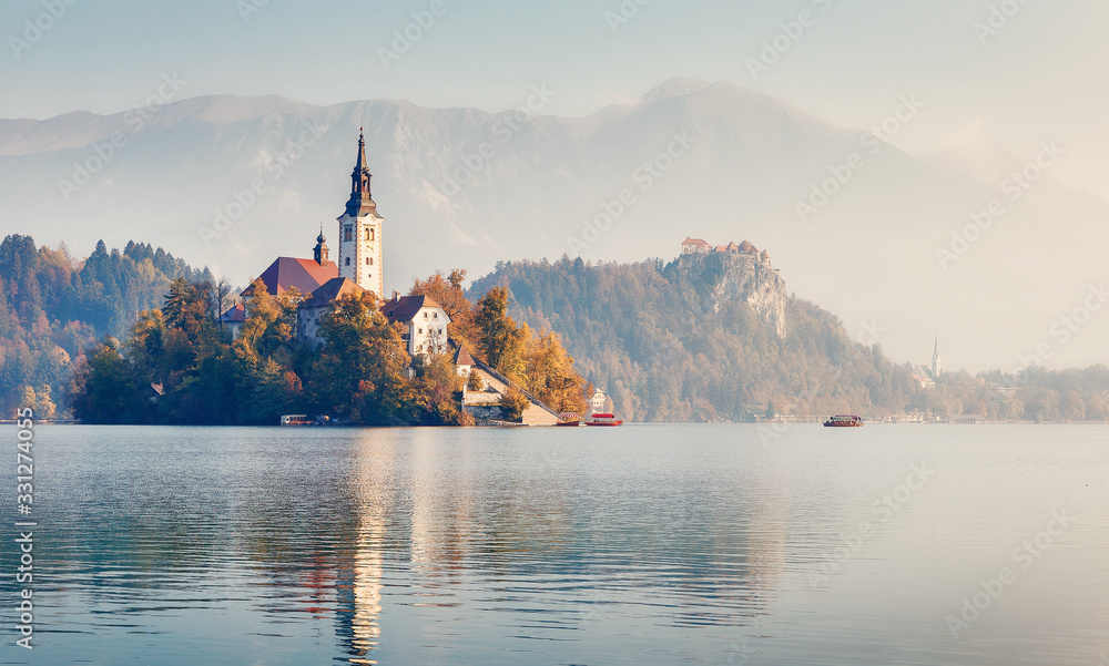Fantastic Sunny morning. Wonderful mountain lake during sunrise. Most famous Slovenian lake and island Bled with Pilgrimage Church of the Assumption of Maria and Bled Castle in background.