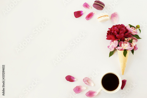 Creative flat lay with ice cream waffle cone with pink and violet peony, petals, coffee cup and macaroons dessert on white background. Beautiful botanical greeting card design, empty space for text