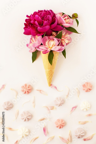 Beautiful fresh peonies in ice cream waffle cone on white background. Frame of pink flower petals and color meringue below. Stylish flat lay with flowers and dessert. Floral card design, copy space.