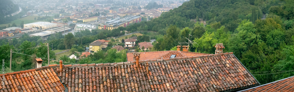 The roofs of the houses are covered with clay tiles. Behind the roofs in the valley, inhabited locality and trees are visible. Horizontal panorama. Background with tiled roofs and the city.