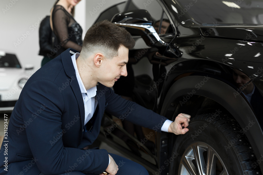 Attractive elegant man examining wheels of a new automobile on sale at dealership. Handsome male driver choosing new car to buy Attractive elegant man examining wheels of a new automobile on sale at dealership. Handsome male driver choosing new car to buy