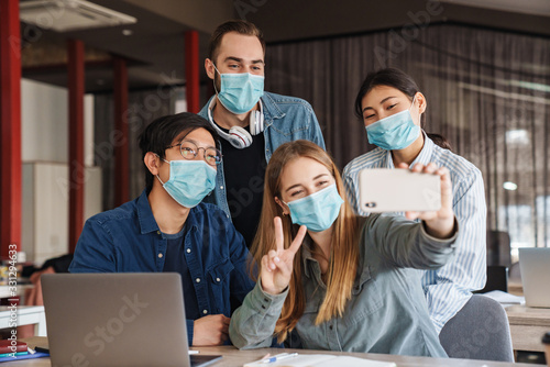 Photo of cheerful students in medical masks taking selfie on cellphone