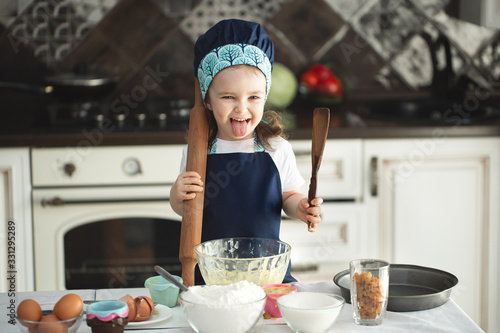 A cute little girl in an apron and a Chef's hat is holding a rolling pin and a wooden spatula, looking at the camera and showing her tongue.