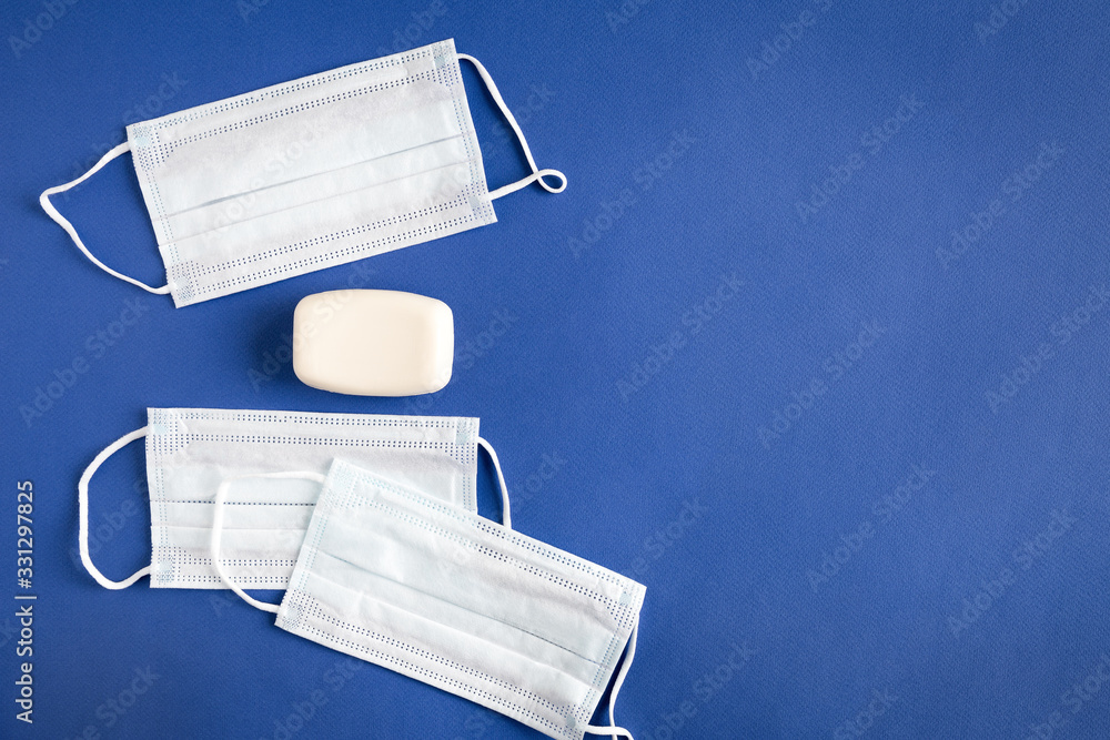 Few isolated objects on a blue paper background with copy space. Three face mask protection against flu, pollution, virus and coronavirus. Next to it is a piece of white hand soap. Few isolated objects on a blue paper background with copy space. Three face mask protection against flu, pollution, virus and coronavirus. Next to it is a piece of white hand soap.