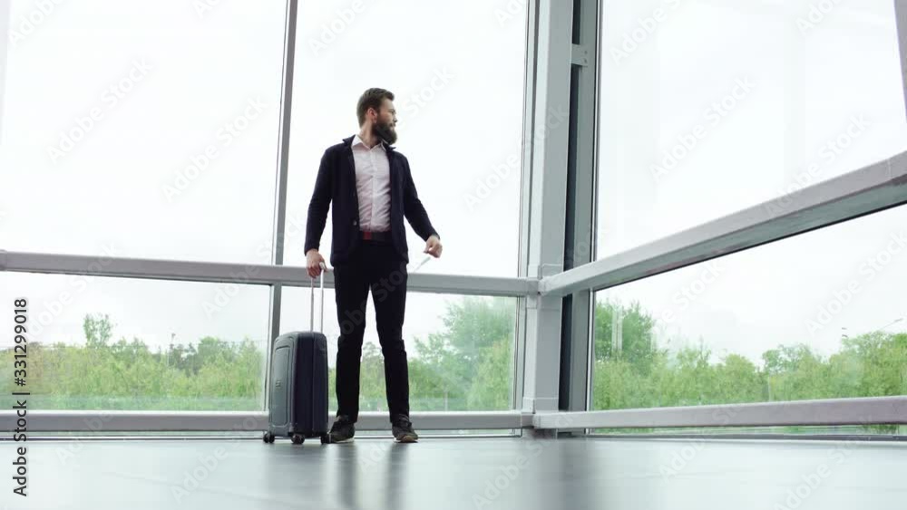 Bearded brunette man in casual clothes is standing with suitcase on wheels near panoramic windows, looking at his watch and waiting for someone. Young guy with luggage is looking forward another Bearded brunette man in casual clothes is standing with suitcase on wheels near panoramic windows, looking at his watch and waiting for someone. Young guy with luggage is looking forward another
