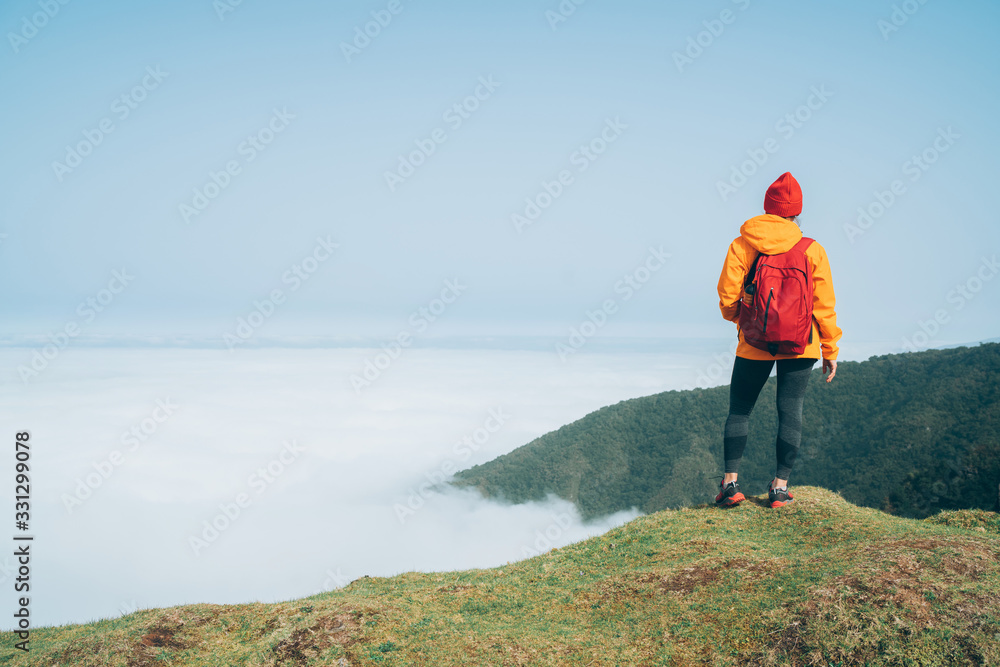 Young female backpacker dressed orange waterproof jacket hiking by the mountain above the cloud route at the end of February on Madeira island, Portugal. Active people around  World traveling concept.