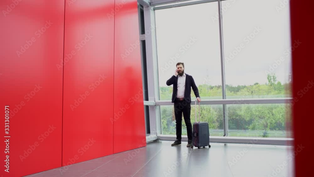 Brunette bearded man is standing with wheeled suitcase, talking on the phone near panoramic windows and then leaving room with red walls. Green trees and busy road outside the window. Brunette bearded man is standing with wheeled suitcase, talking on the phone near panoramic windows and then leaving room with red walls. Green trees and busy road outside the window.