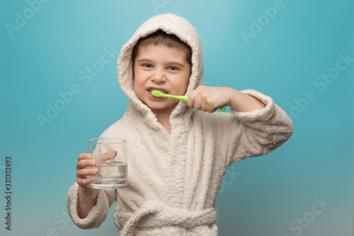 the boy brushes his teeth. A child in a robe with a toothbrush and a glass of water on a blue background.