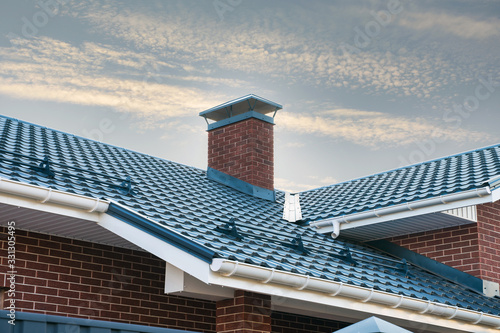 Newly installed construction frame house rain gutter system on the metal roof top. Brick chimney pipe against blue sky.