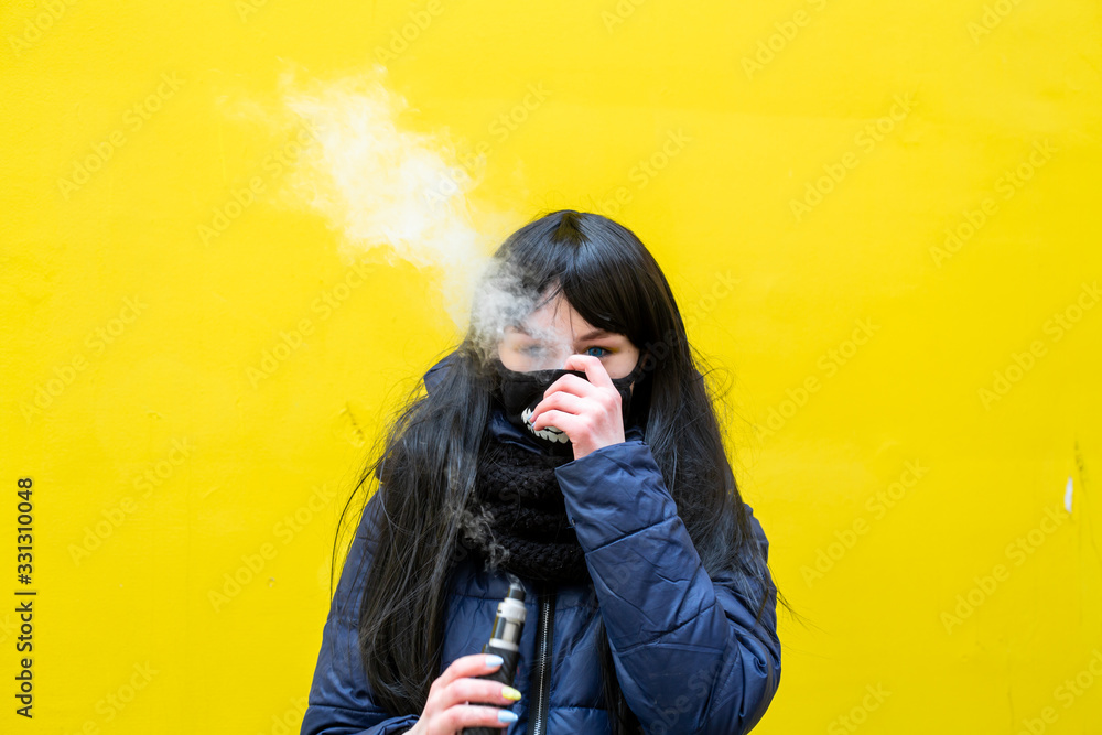 Obraz premium Vape teenager. Young pretty white caucasian brunette girl with blue eyes and in a blue jacket smoking an electronic cigarette opposite yellow wall on the street in spring. Virus protection. Bad habit.