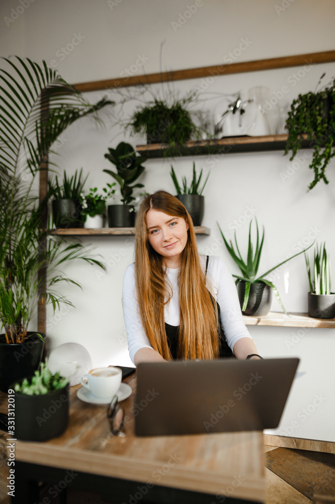 Cute freelancer girl with laptop working in a cozy cafe, looking into the camera and smiling. Attractive student girl uses the internet on a laptop in a stylish coffee shop. Vertical
