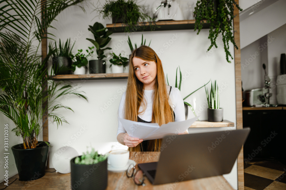 Funny lady with a laptop on the table and business papers in her hands, looking away with a pensive face on a white wall with plants. Freelancer girl working with laptop and documents in coffee shop Funny lady with a laptop on the table and business papers in her hands, looking away with a pensive face on a white wall with plants. Freelancer girl working with laptop and documents in coffee shop
