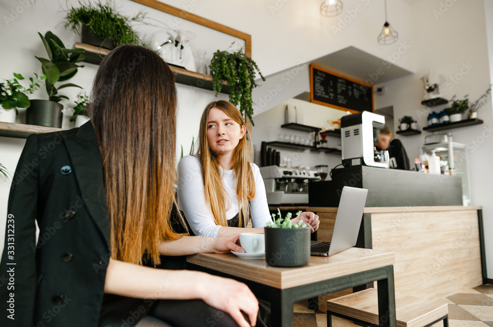 Two attractive young businesswomen , sitting in a light cozy cafe with a laptop, drinking coffee and discussing a joint business project. Business meeting of two young stylish girls in a coffee shop. Two attractive young businesswomen , sitting in a light cozy cafe with a laptop, drinking coffee and discussing a joint business project. Business meeting of two young stylish girls in a coffee shop.
