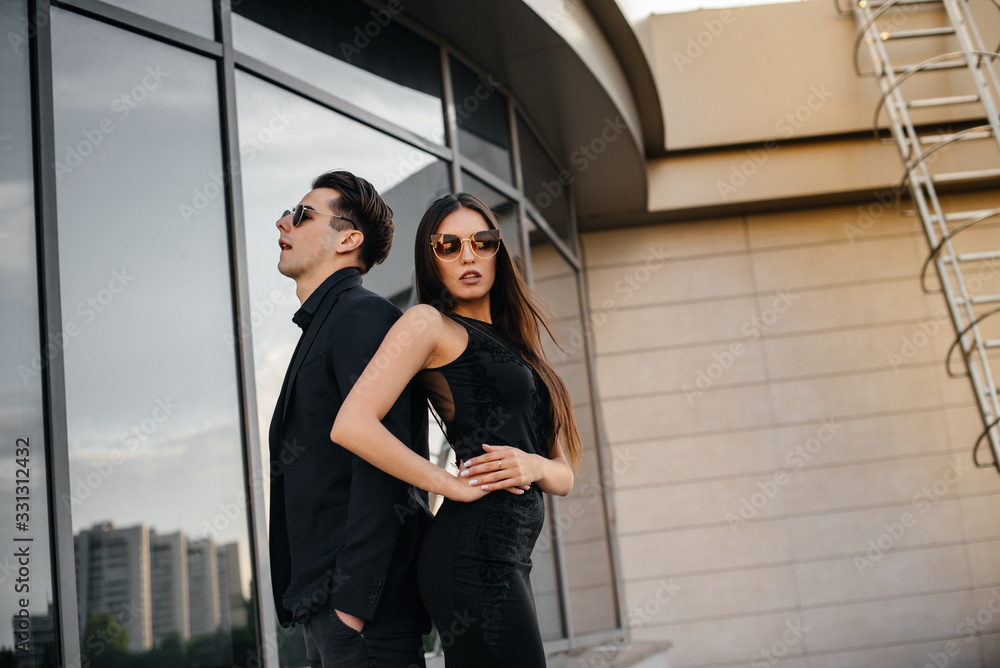 A beautiful, stylish pair of young people in black clothes and glasses stand against the background of an office building in the sunset. Fashion and style