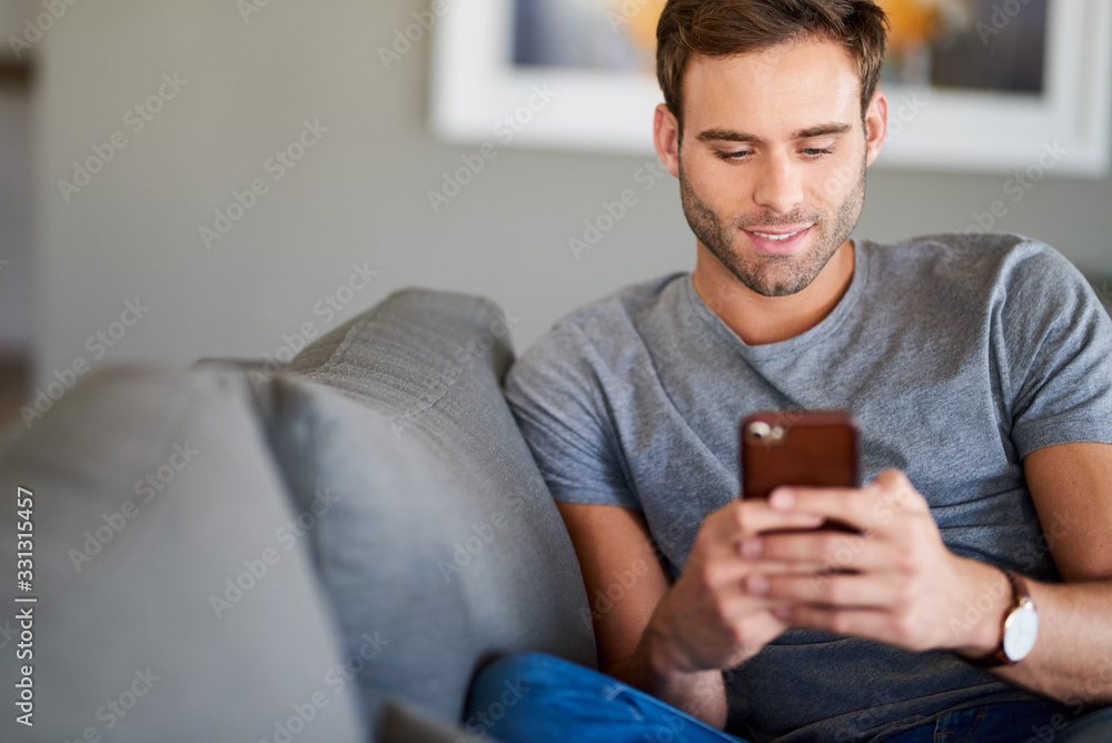 Smiling young man texting on his living room sofa - obrazy, fototapety, plakaty 