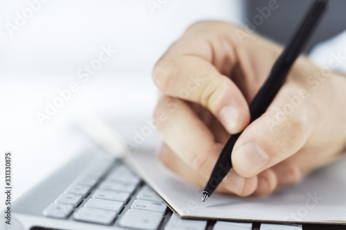 Man writes with a pen in notepad on computer keyboard in a sunny office, business and education concept. Close up