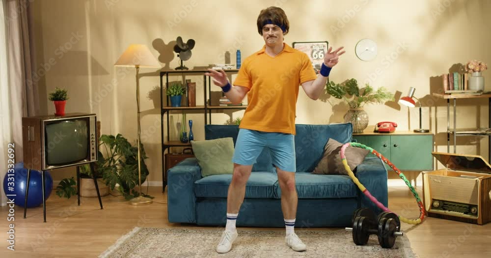 Handsome funny Caucasian retro style man with mustache dancing at blue sofa in room with hula hoop at home. Joyful smiling athlete male model fooling around while posing to camera indoors. 50s concept