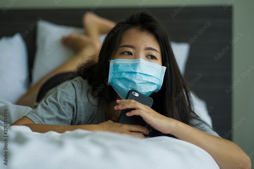 pandemic and virus outbreak - young beautiful scared and worried Asian Korean woman in medical mask checking online news with mobile phone on bed lockdown at home pandemic and virus outbreak - young beautiful scared and worried Asian Korean woman in medical mask checking online news with mobile phone on bed lockdown at home
