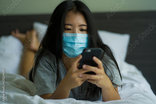 pandemic and virus outbreak - young beautiful scared and worried Asian Korean woman in medical mask checking online news with mobile phone on bed lockdown at home