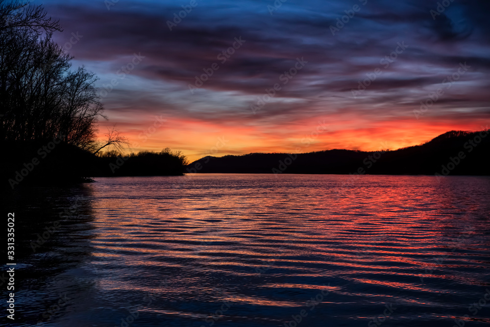 The sunset paints the sky over the hills of the Ohio River Valley with beautiful color as reflections dance on the surface water ripples. Ohio is viewed from the West Virginia river bank.