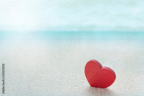 Red wooden heart model on the sandy sea beach with blue sea blurred background.