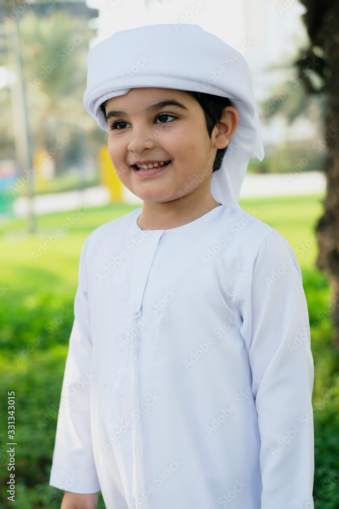 Young Emirati Boy wearing traditional Kandura disdash for kids while at an outdoor park with grass, trees and natural sunlight as background Young Emirati Boy wearing traditional Kandura disdash for kids while at an outdoor park with grass, trees and natural sunlight as background