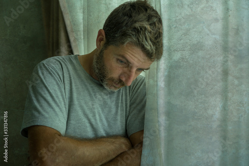mid adult man crying sad and depressed standing at bedroom window in despair at home feeling worried and overwhelmed suffering depression and anxiety problem