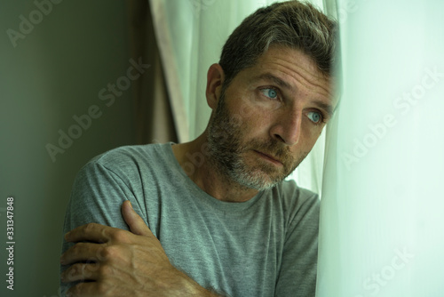 mid adult man crying sad and depressed standing at bedroom window in despair at home feeling worried and overwhelmed suffering depression and anxiety problem