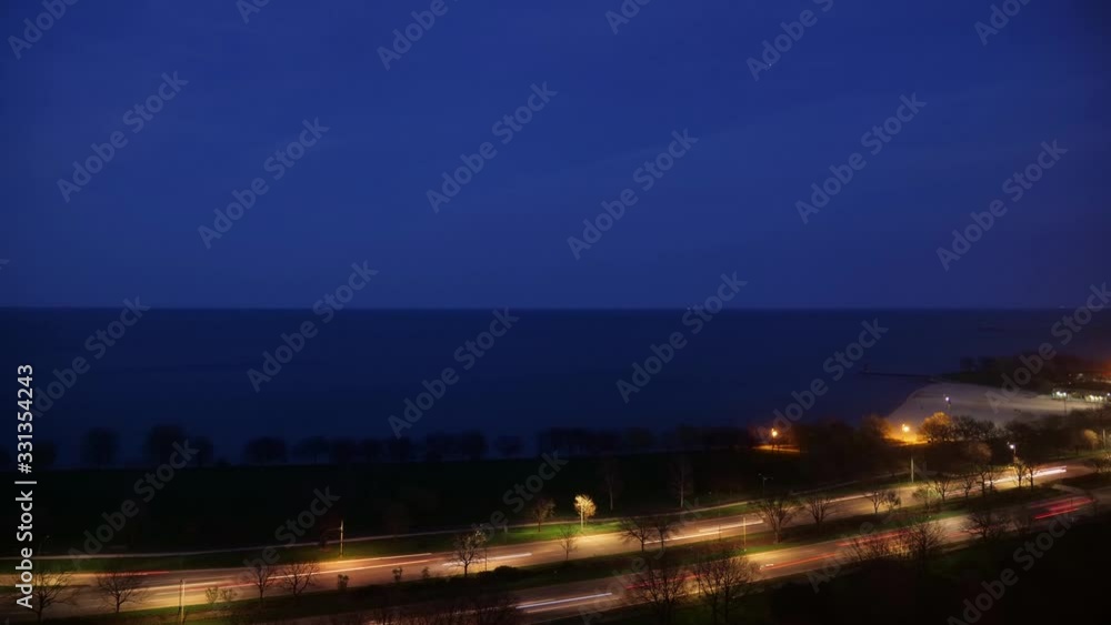 Timelapse of vehicle traffic light trails on the busy Lake Shore Drive at night as the lights from airplanes of fly overhead creating copy space in dark blue night sky over Lake Michigan.