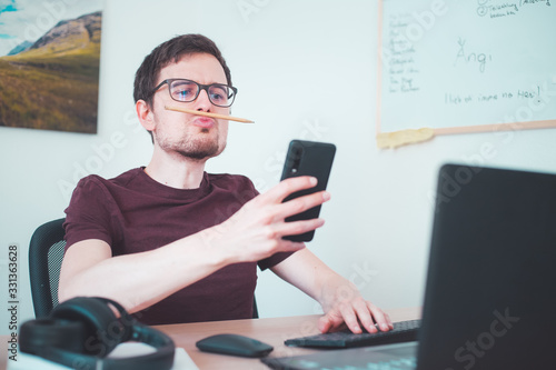 Bored freelancer concept. Young man is fooling around on his workplace with a pencil trapped between nose and lips photo