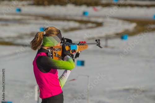 Female biathlon racer is standing on the ground and aiming her rifle. Biathlete woman on a shooting range, firing standing up. photo