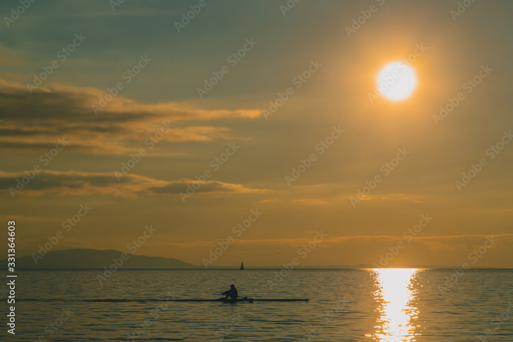 Kayaker or a person in a paddle boat on a lake during evening hours. Romantic leisure activity on the water. Sun about to set into the water.