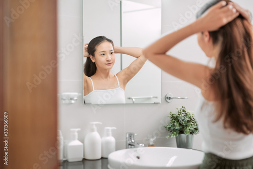 Young redhead woman tying her hair up in a bun in front of the mirror in a bathroom viewed from the rear