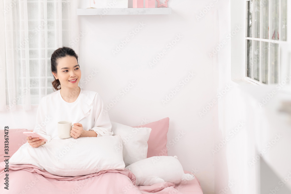 Woman in bed in the bedroom with a mug and a phone in her hands Woman in bed in the bedroom with a mug and a phone in her hands